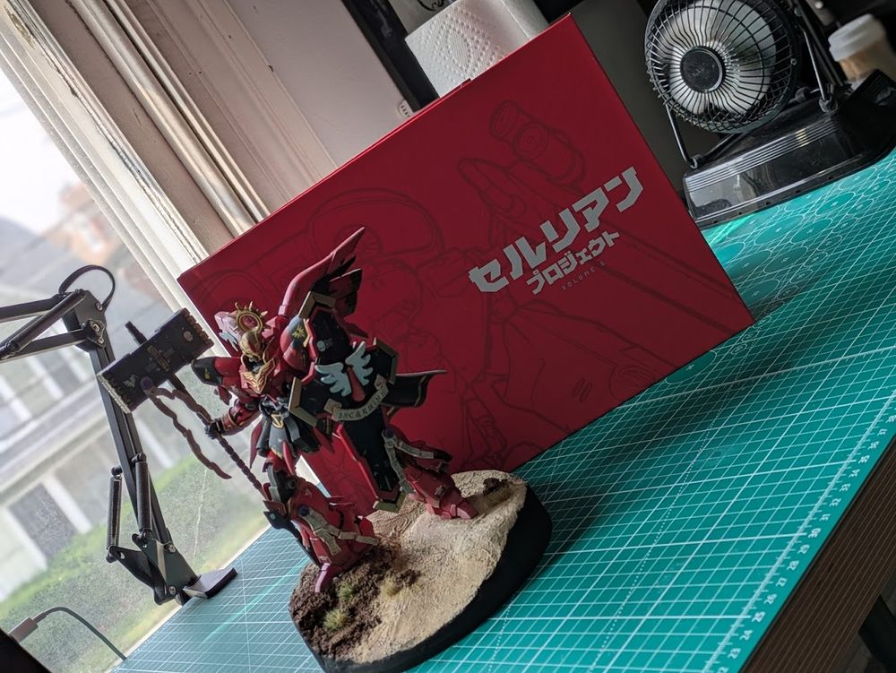 A large red landscape format hardcover book sitting on a desk covered by a large green cutting mat. In front of the book stands a customized gunpla figure with a hammer and shield, it is also painted red.