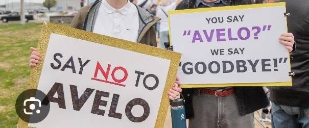 Two protestors hold signs. One signs says “SAY NO TO AVELO” and the other says “You say AVELO? We say GOODBYE!”