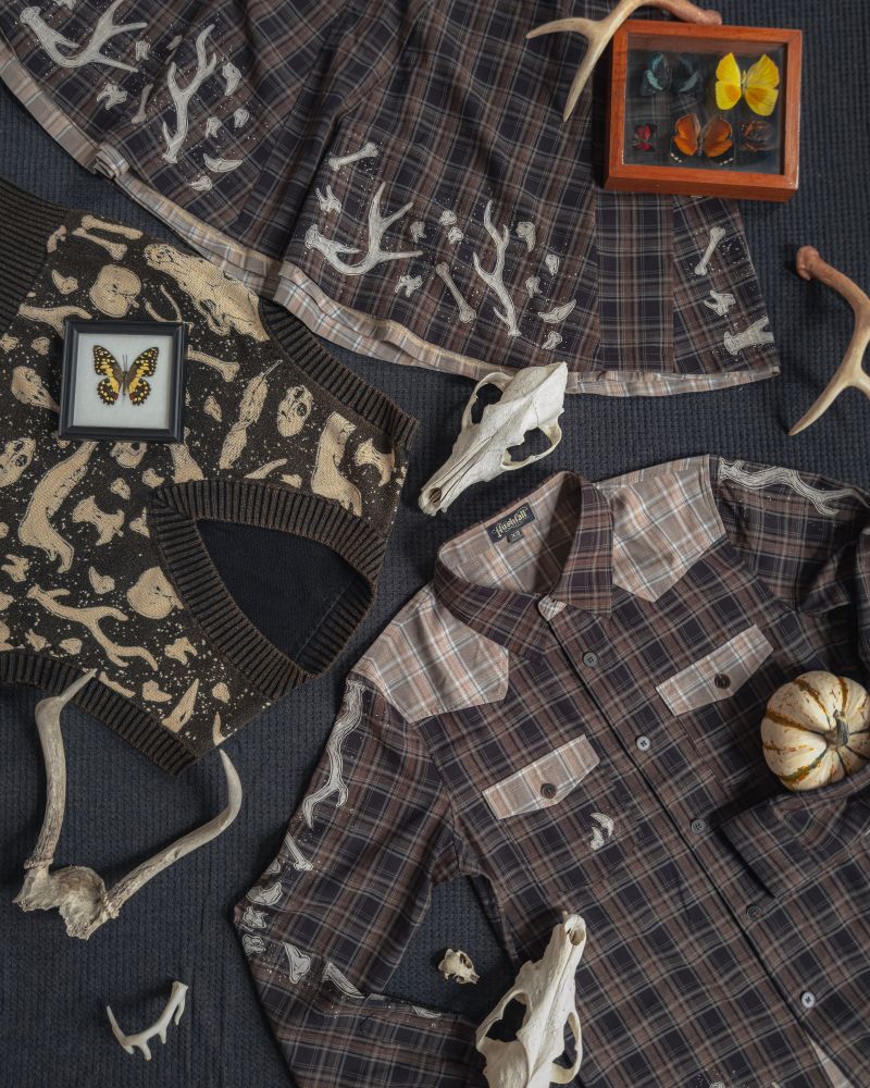 a still photo of a warm black-and-white Decomposition Vest, the Bone Collector plaid skirt, and Bone Collector plaid shirt among a smattering of bones and pinned butterflies. All designs by the Hushfall Archives :) 