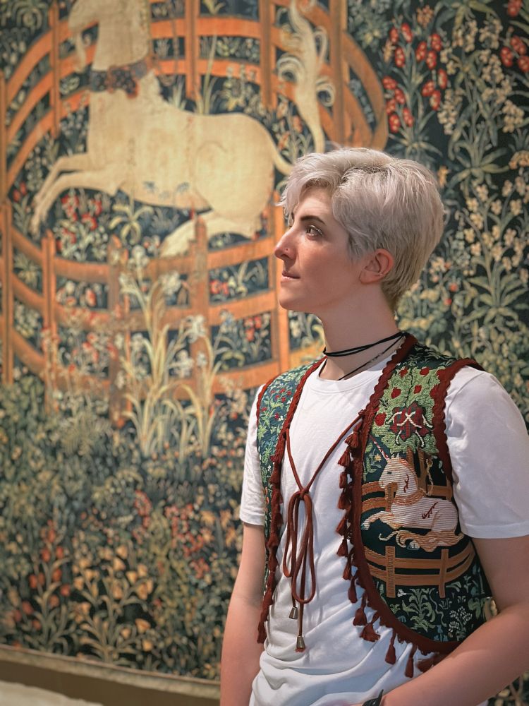 a still image of Emmett, the Archivist, wearing our Unicorn in Captivity inspired vest. blue brocade with green leaves showcases a unicorn underneath a pomegranate tree, surrounded by a gold fence. red cotton fringe adorns the vest, and long brass-capped ties close the vest in front. 