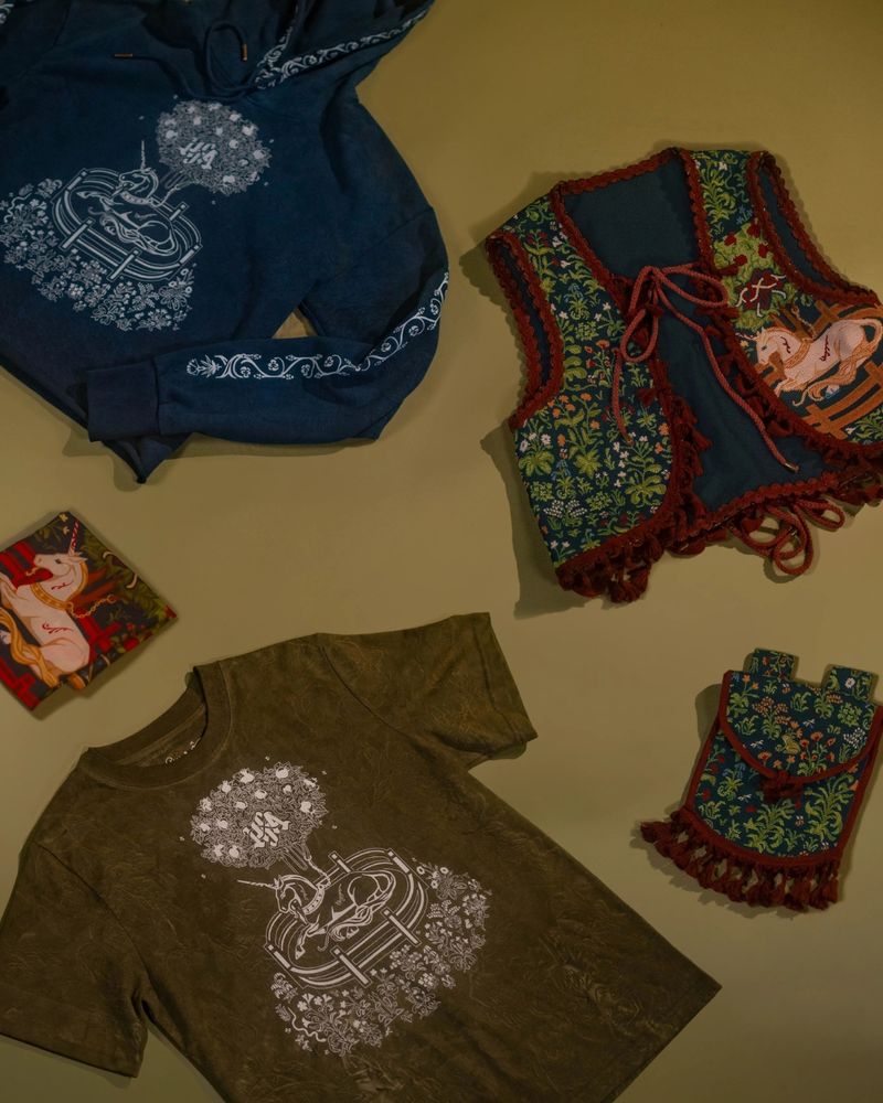 A still image with a mossy green background, showcasing clothing items inspired by the Unicorn In Captivity, a 15th century tapestry. 