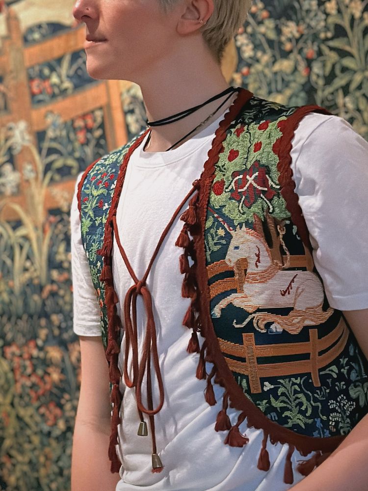 a still image of Emmett, the Archivist, wearing our Unicorn in Captivity inspired vest in a close-up crop. blue brocade with green leaves showcases a unicorn underneath a pomegranate tree, surrounded by a gold fence. red cotton fringe adorns the vest, and long brass-capped ties close the vest in front. 