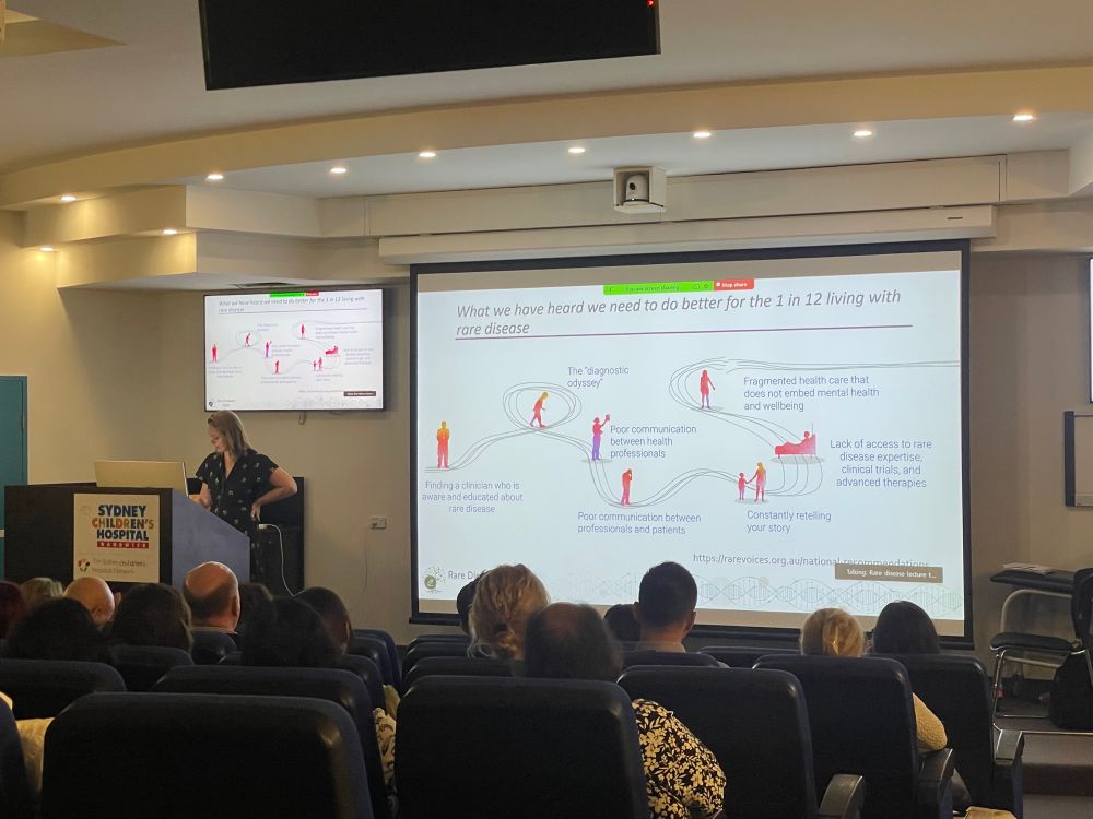 Lecture theatre with woman speaking at a podium at the front and a large projection screen. 