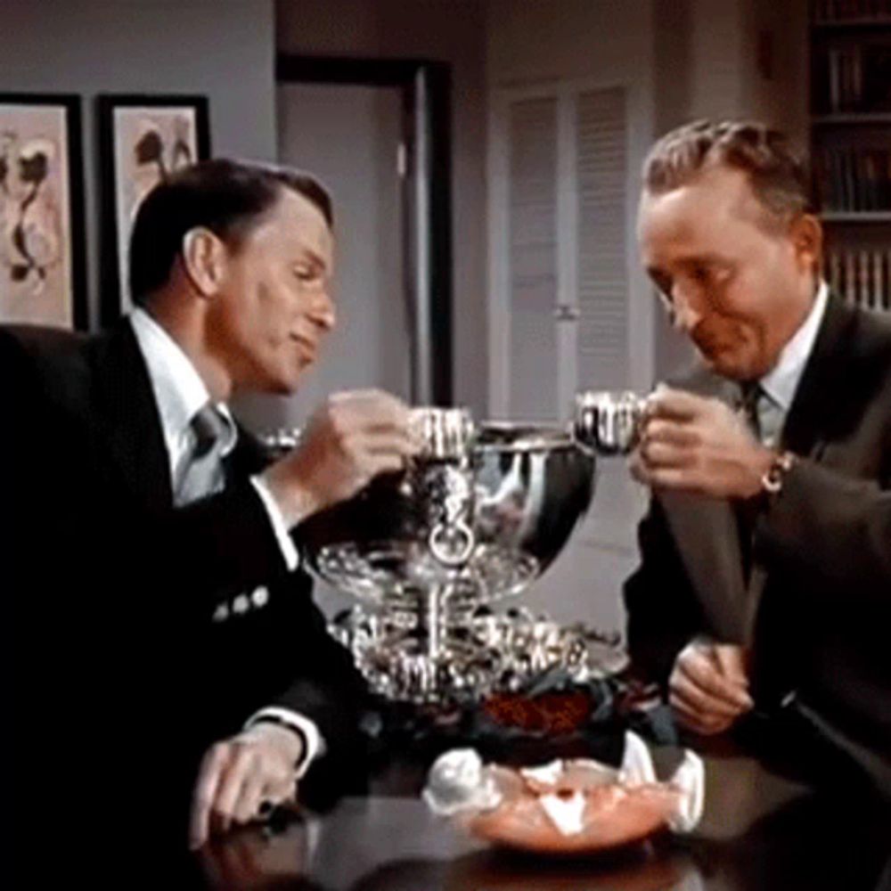 two men in suits are sitting at a table toasting with cups .