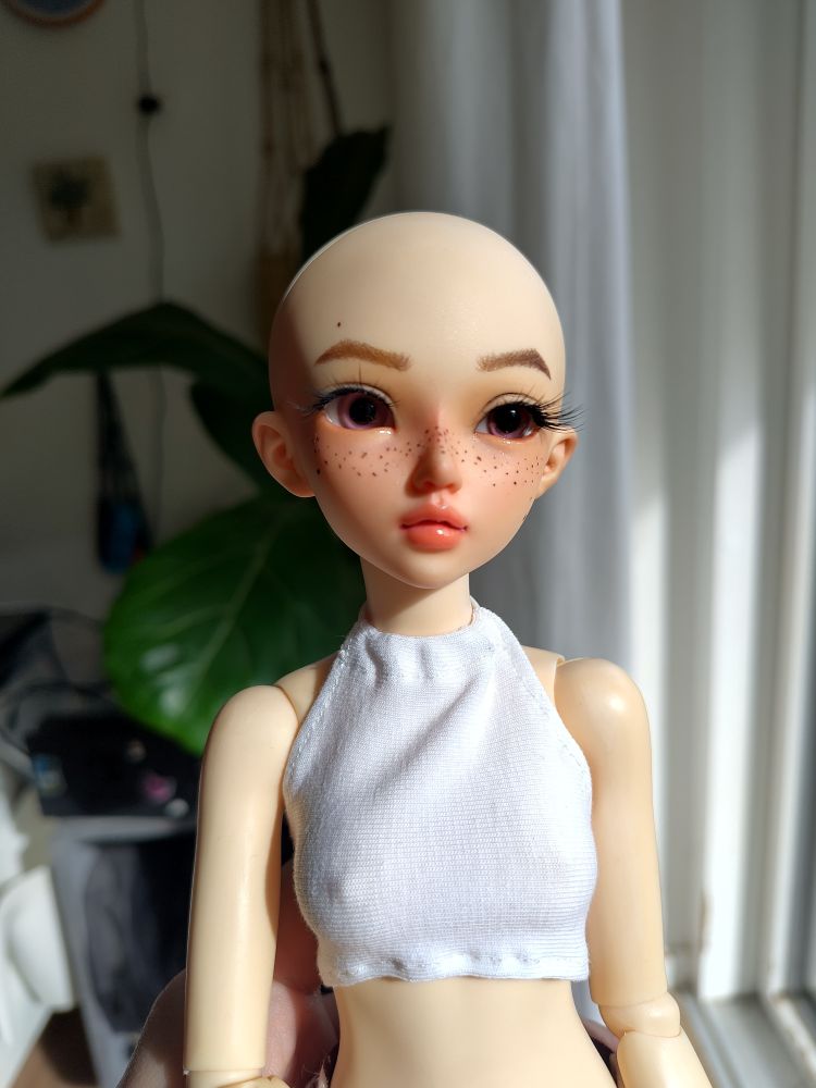 Bald bjd held up by me, she's wearing a white crop top, she has pink/purple eyes and freckles 