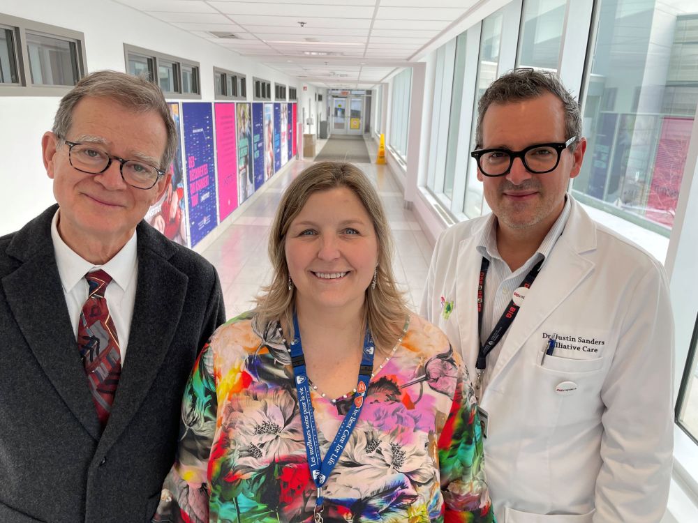 Un homme port des lunettes et un costume noir; une femme porte une chemise vibrante; un homme port des lunettes et un manteau de médecin. Ils posent dans un couloir.