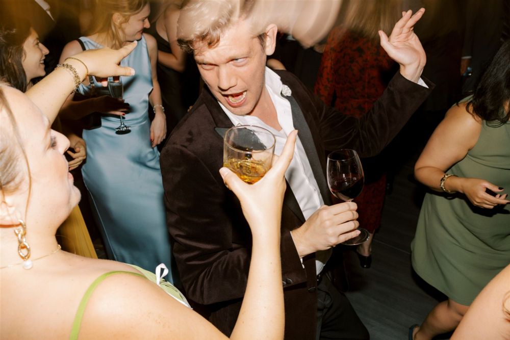 Brady dancing on a crowded wedding dancefloor, holding a glass of red wine 