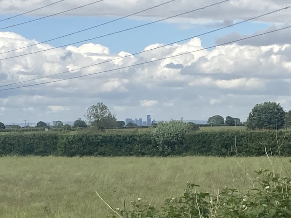 At extreme zoom across the fields, the skyscrapers of Manchester City centre.