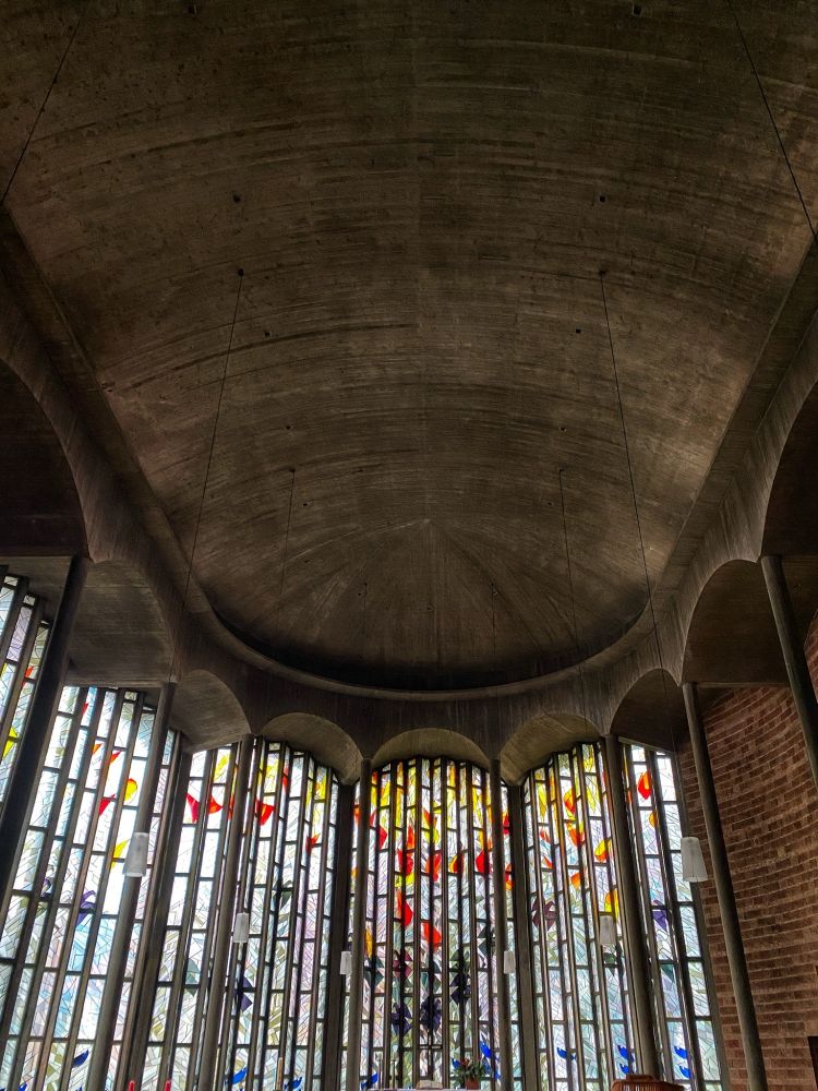 Photograph of the Ludwigskirche ceiling. It's concrete, oblon,g and rounded at the end. Besides the ceiling, the photo shows the wall behind the altar - most of it covered in stained glass work, piling up with rectangular vertical pieces. It's abstract and mostly light, featuring yellows and reds in the upper areas and few darker shades of blue and green lower to the bottom. It doesn't cover the whole wall symmetrically, segments on the right are a wall made out of red, unplastered bricks.