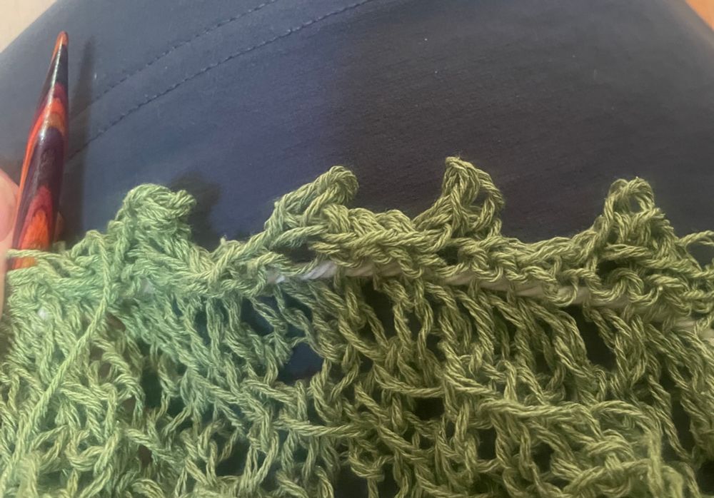 Edge of bound-off green silk yarn knitting project - picot edge is 4 little bumps. Laying against dark blue skirt. Orangish needle left of frame.