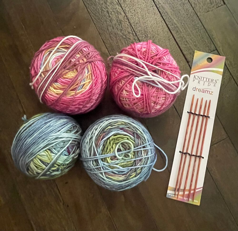 Four cakes of yarn, two pinkish and two blue/greenish - self-striping skeins for socks. Beside a set of really small US1 double-pointed needles in packaging. On dark brown table.
