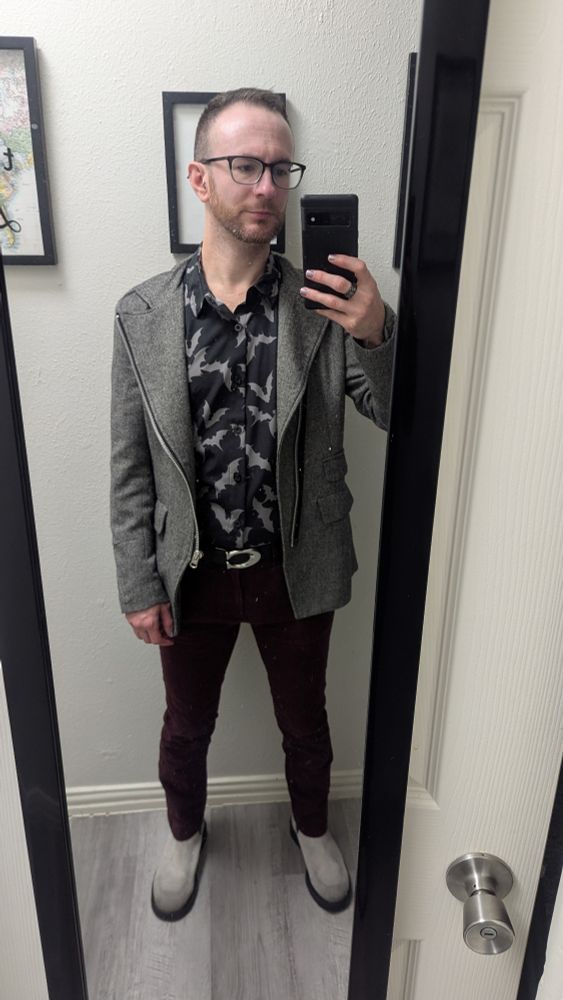 A faggot in a black button front shirt with gray bats on it, maroon skinny khaki pants, gray suede Chelsea boots, and a gray herringbone blazer with asymmetrical zip front.