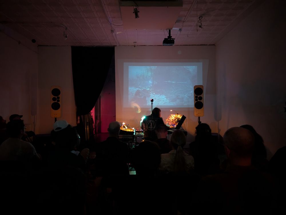 dimly lit room. Alex and modular synth setup in the middle. my 3-way panel hifi speakers in the corners. video of cave diving playing overhead