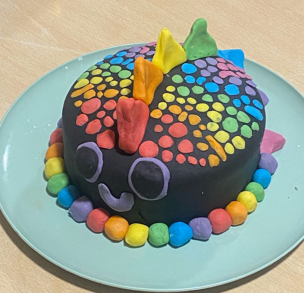 A cake covered with black fondant and colourful decorations to somewhat resemble a rainbow dinosaur