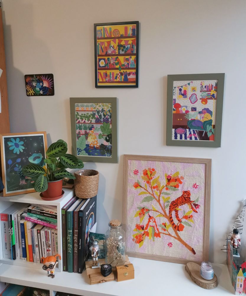 Photo du mur au dessus de ma bibliothèque Kallax. On y voit 3 print de Marie Boiseau accrochés au mur, un Canevas Fatal et une 4ème print posés sur la bibliothèque. On voit aussi une petite maranta et des livres. 