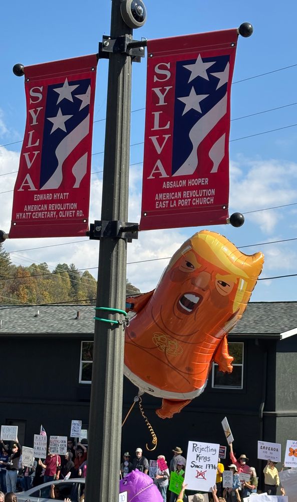 A balloon of a whiny, diapered Trump baby with man boobs flies next to flags celebrating Revolutionary War heroes 