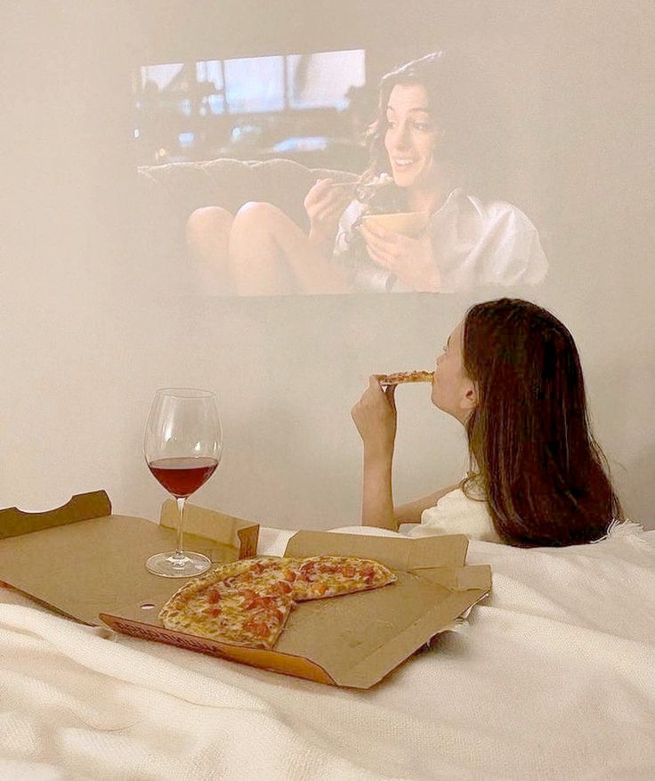 [picture of a woman with brown hair watching a movie being projected on her bedroom wall. the woman is eating pizza straight out of the box, and there is a glass of wine sitting on the box as well. the movie is an anne hathaway joint, bc taste.]