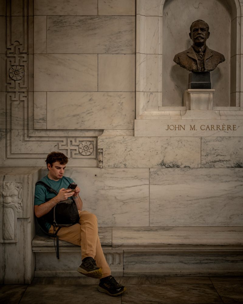 A white man with red hair sits on a bench in the New York public library. The marble wall and bench are a neutral background to his orange pants and teal shirt. He is on his phone. In the upper right corner of the image is a bust of one of the architects of the library - John M Carrere.
