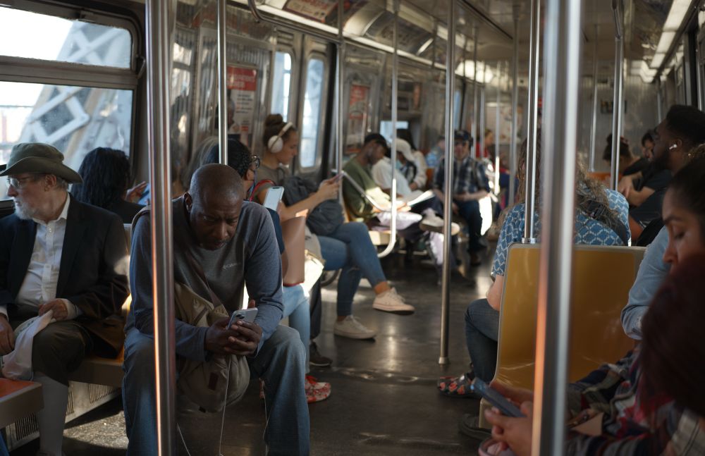 A Black man in a grey long sleeve shirt is looking at his iPhone on the Q train. A white man wearing a green boonie hat and black sports coat sits next to him looking out of the window. In the background are various subway riders. In the foreground a woman is reading an article on her phone.
