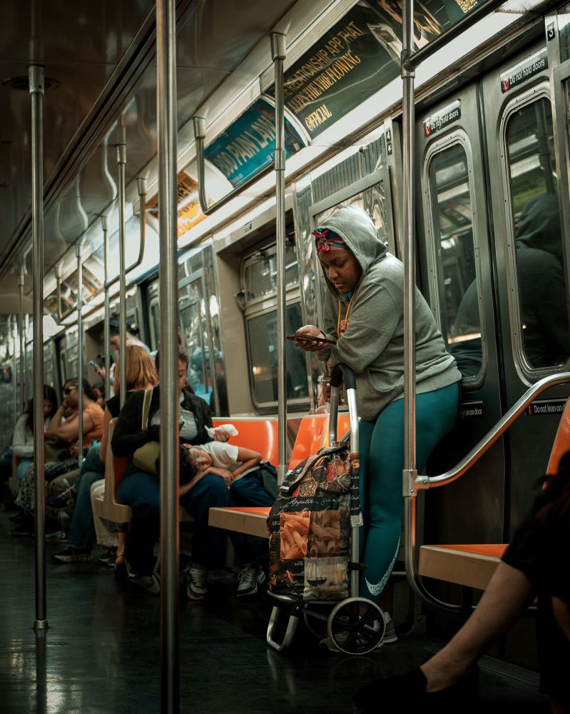 On the Q train a Black woman leans against her shopping trolley. She’s wearing teal pants and a gray hoodie while looking at her phone. In the background a boy is seen laying in his caretakers lap against the contrasting orange seats of the train.