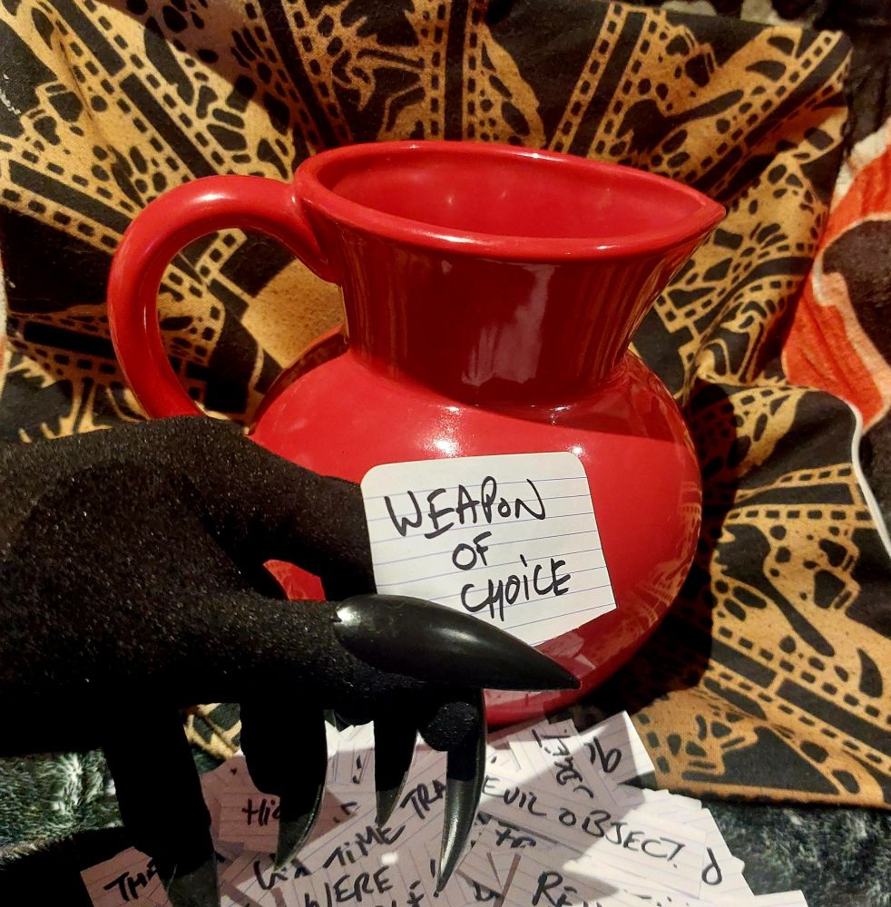A clawed hand holds a piece of paper that reads, 'weapon of choice'. In the background is a large bold red ceramic jug, with a pile of similar prompts written on slips of paper. 