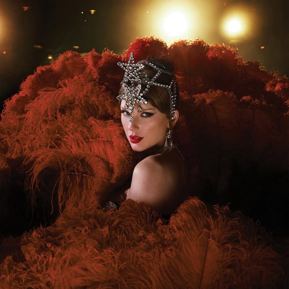 Taylor Swift is staring at the camera. She has on a headpiece with two bedazzled stars on it. She is surrounded by maroon feathers. There are stage lights behind her