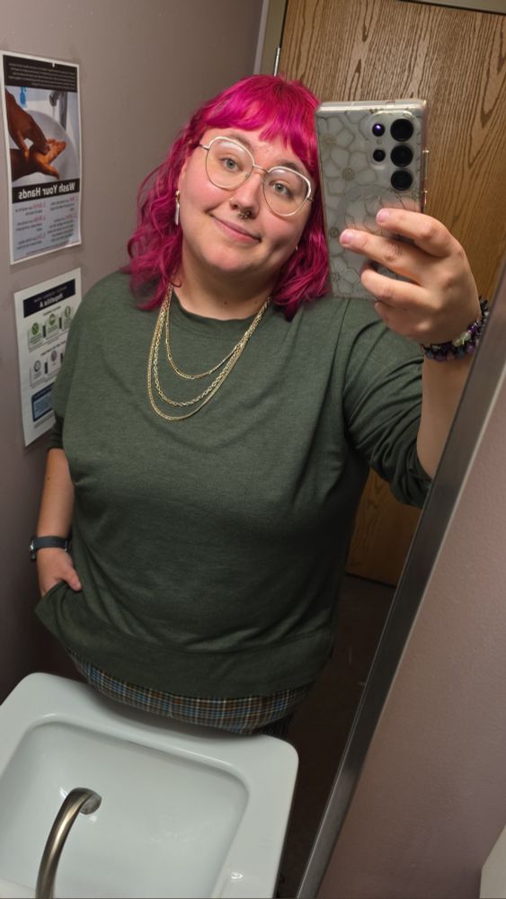 A person takes a mirror selfie. They have bright pink hair. They are wearing a green sweater, checked trousers, gold chains, and ghost earrings.