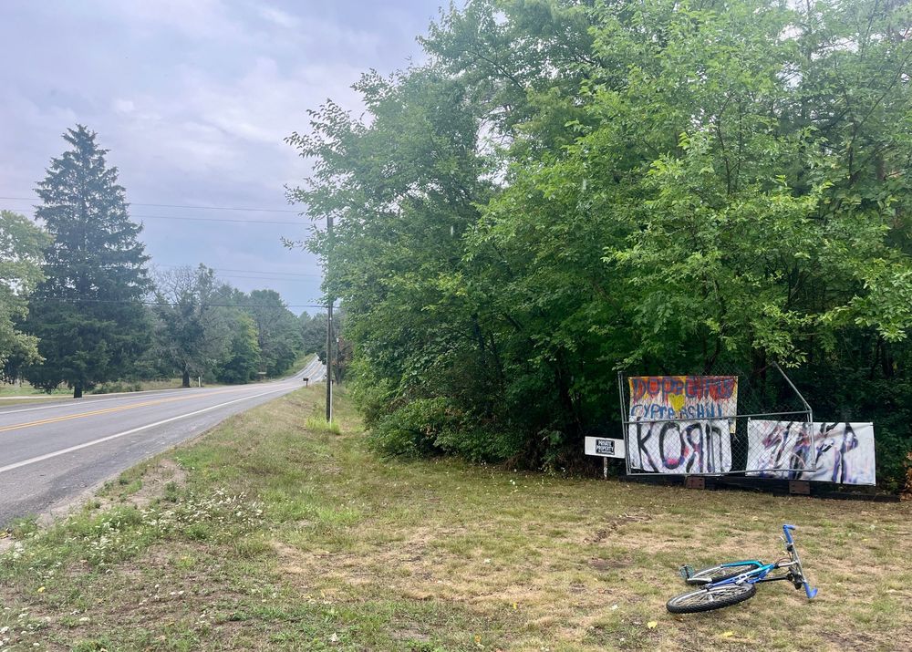 A random roadside in rural West Michigan, with woods, hand painted metal band logos, no trespassing sign, storm clouds, and a blue bike
