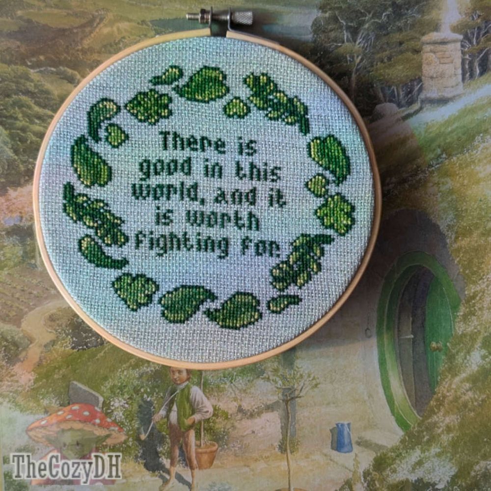 A cross-stitched piece framed in a hoop with the quote 'There is good in this world, and it is worth fighting for' surrounded by leaves. The hoop rests on an Alan Lee painting of the Shire.