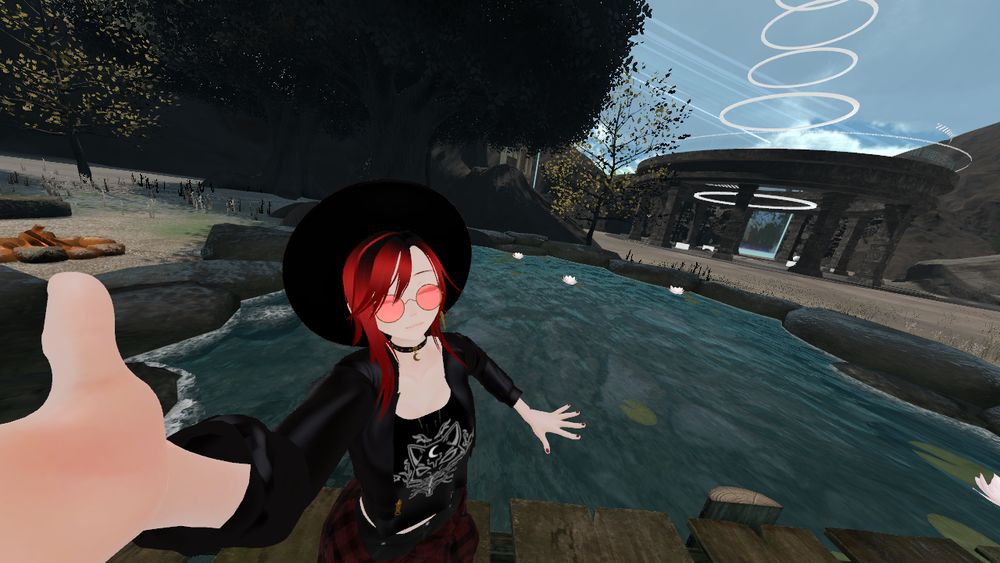 Photo shows me taking a selfie with my avatar in the VRChat world - The Museum of Immersivism by kgpaints. I am standing on a virtual bridge, over a pond with water lillies. There are trees and a campfire in the background. There is also a an ancient ruin like structure with circles of light shooting up from the middle, a bit like a rings of a Sci-fi teleporter. 