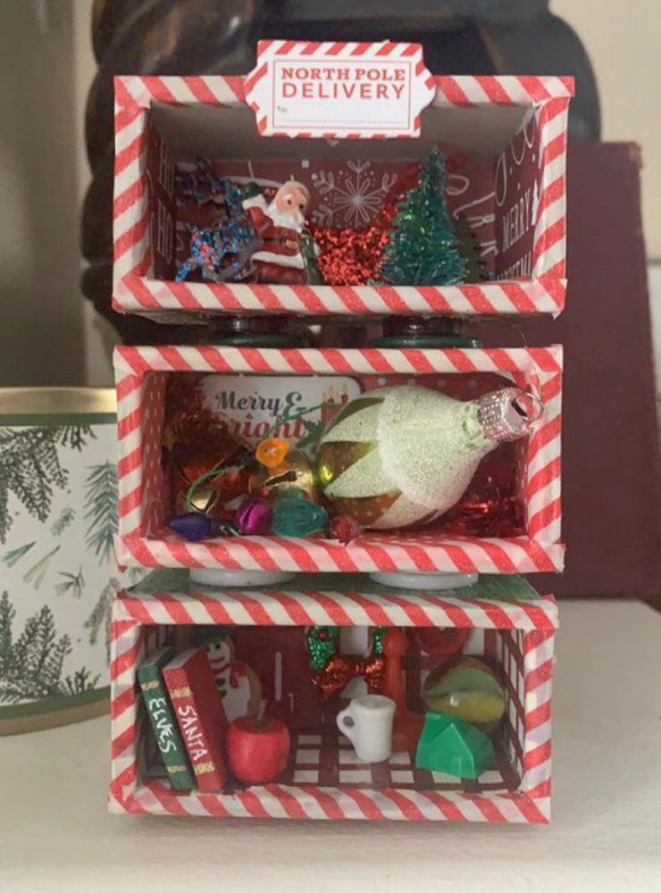 3 boxes stacked- ea box 3x2x2” buttons in between boxes to separate them. Ea box covered in red/white striped paper 
Ie.bottom box 
Two books leaning on left “wall” one labeled Santa, the other Elves. A gingerbread cookie snowman leaning on back wall, a coffee mug, a little house from Monoply, a marble, a wreath on back wall.
