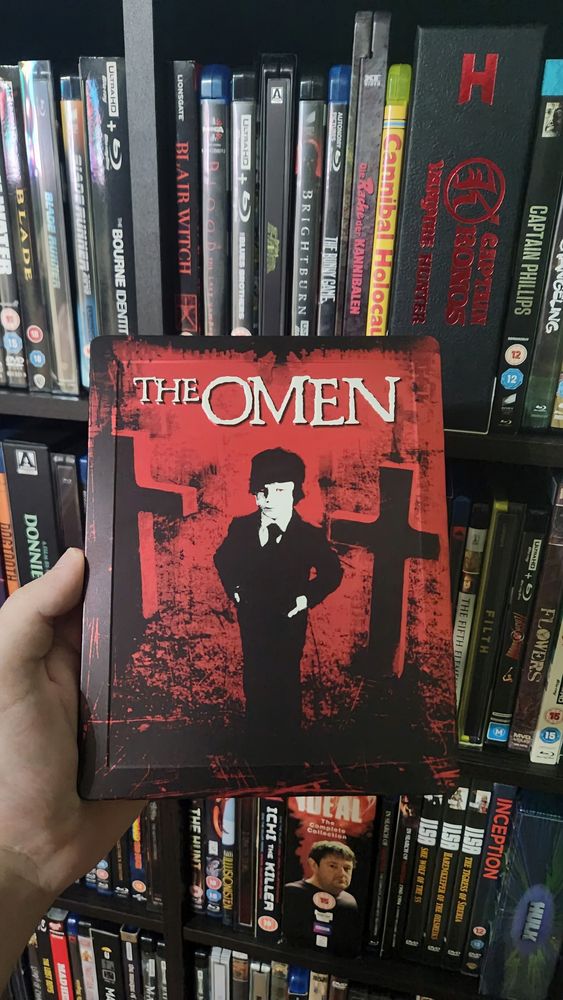 A steelbook being held in front of a movie collection. The artwork is an illustration of a child standing next to two gravestone crosses. The background has a red filter and the child's eyes are also red. At the top is the movie's title "The Omen"