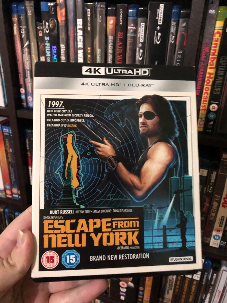 4K blu ray cover featuring a muscular man with an eyepatch holding a rifle with a scope on it. In the background is a geographical graphic of New York City with “Escape From New York” written in yellow text in the lower left hand corner 