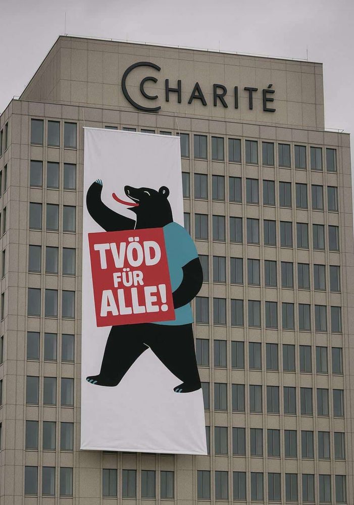Foto des Bettenhochhauses der Charité in Berlin Mitte.
Daran befestigt ein großes Fall-Banner mit einem abgewandeltenBerliner Bären der ein Schild trägt auf dem "TVÖD FÜR ALLE!" zu lesen ist.
