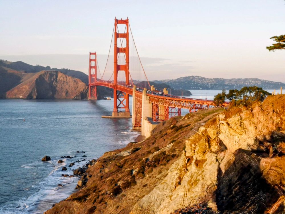 A stunning landscape shot of the Golden Gate Bridge at sunset, viewed from the rugged, rocky coastline of the Marin Headlands. The bridge's iconic International Orange towers and roadway stretch across the blue water, with traffic visible on the deck. The foreground is dominated by sun-kissed, golden-brown cliffs and a rocky beach where waves crash gently. In the distance, the hills of San Francisco are visible, hazy in the late afternoon light. The sky is a pale blue, with the warm glow of the setting sun illuminating the bridge and the hillside.