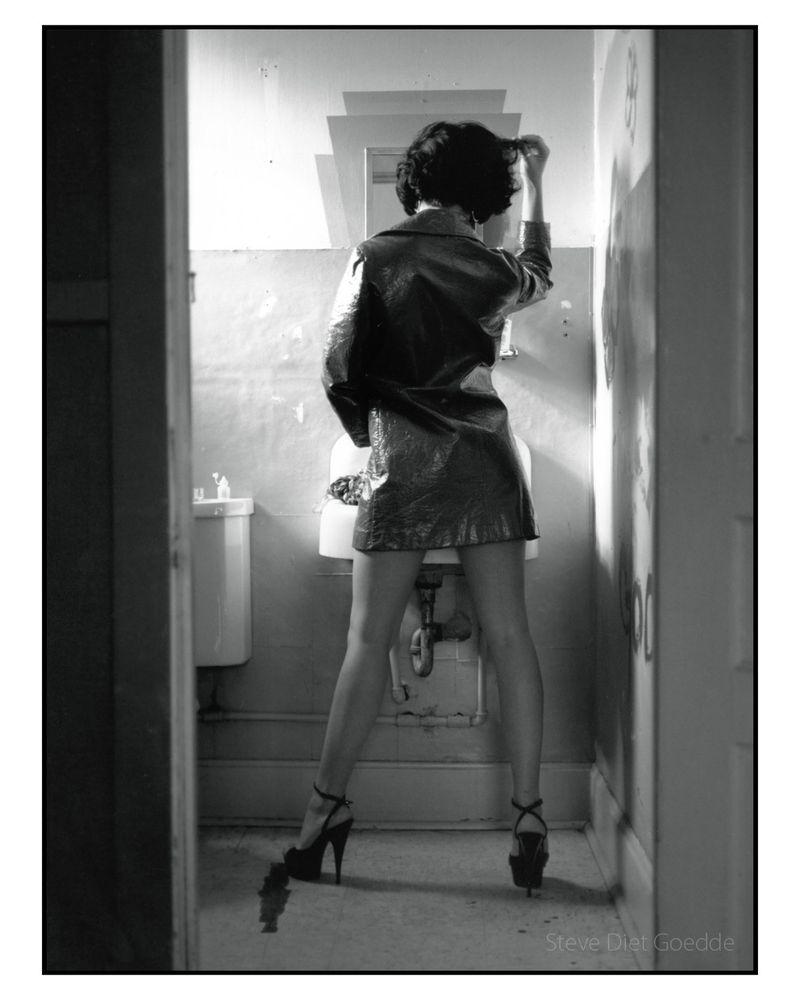 A black and white photograph of a model with her back to the camera standing in front of a bathroom mirror seemingly inspecting her makeup. She is wearing a shirt vinyl coat revealing her long legs. She is also wearing high heeled platform shoes. #legs #highheels #heels #platforms #leggy #vinylcoat