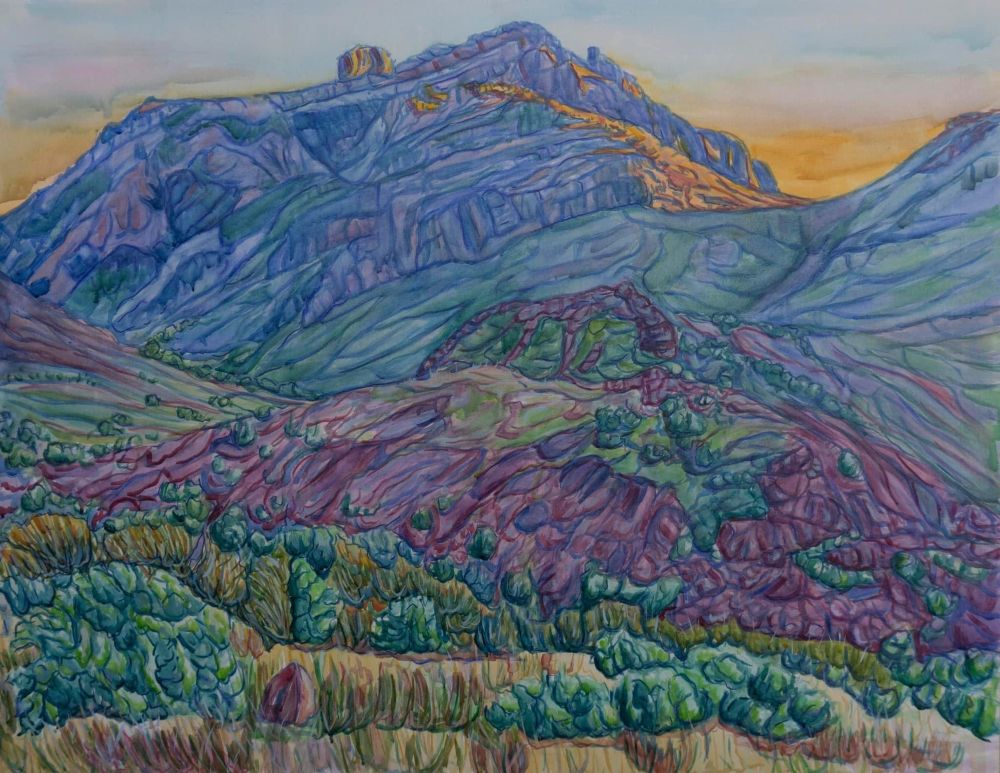 Blue & purple shaded hills & peaks with green sagebrush bushes, lit up in subtle orange at the top as the sun sets behind them. 