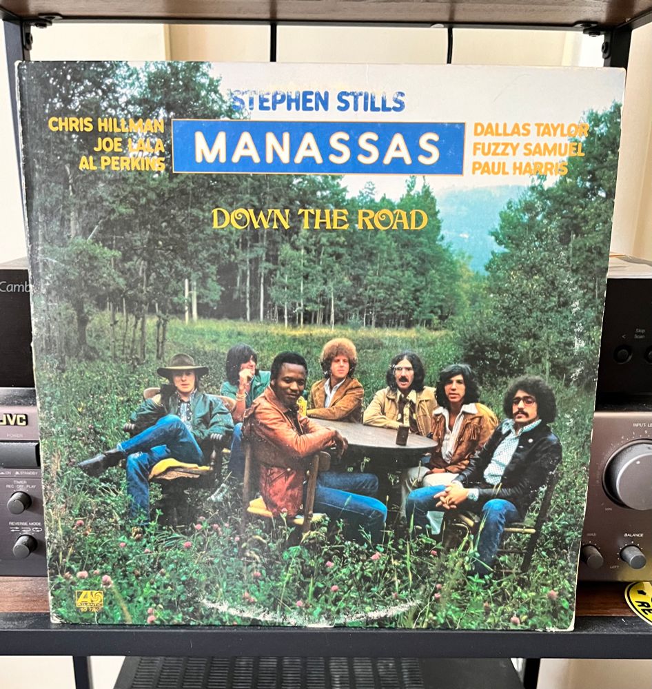 Album cover for ‘Down the Road’ by Manassas. Album cover is a bunch of 70s looking dudes around a table in an overgrown field. Stephen Stills is slightly off to the left. 