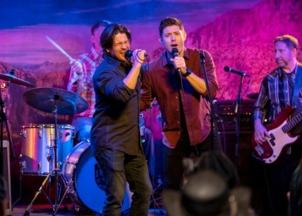 jensen ackles as dean winchester in supernatural season 15 episode 7 last call. he's holding a microphone and has an arm around the actor playing lee webb. the backdrop is lit up like the bisexual flag