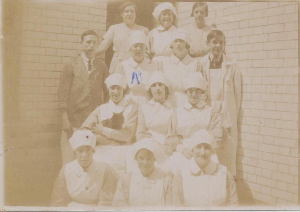 SBHX8/1788 - Thirteen members of the catering staff at St Bartholomew's Hospital, 1922. The eleven women and two men are pictured in four rows. The women are wearing white aprons, collars and caps, and one is holding a black cat. One woman has been marked with the letter M or by an X.