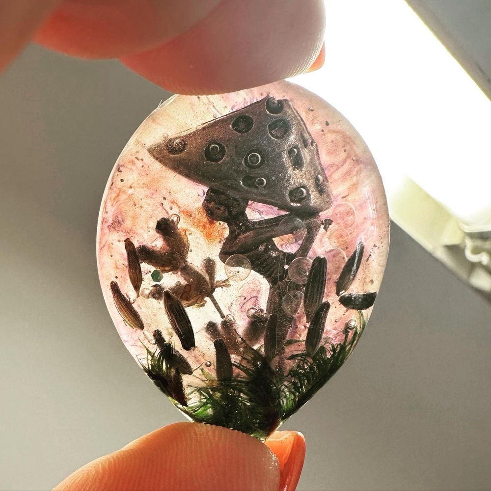 A teardrop shaped pendant with a mushroom faerie, lavender buds, and green glass held up to light to see transparency