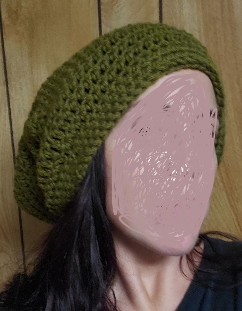 A women with dark hair is standing in front of wood paneling wearing an olive green slouch beanie. Her face is blurred out. 