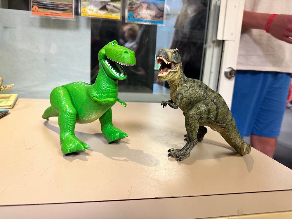 A cartoonish T-Rex doll (Rex from Toy Story) stands next to a realistic one from the Jurassic Park movies