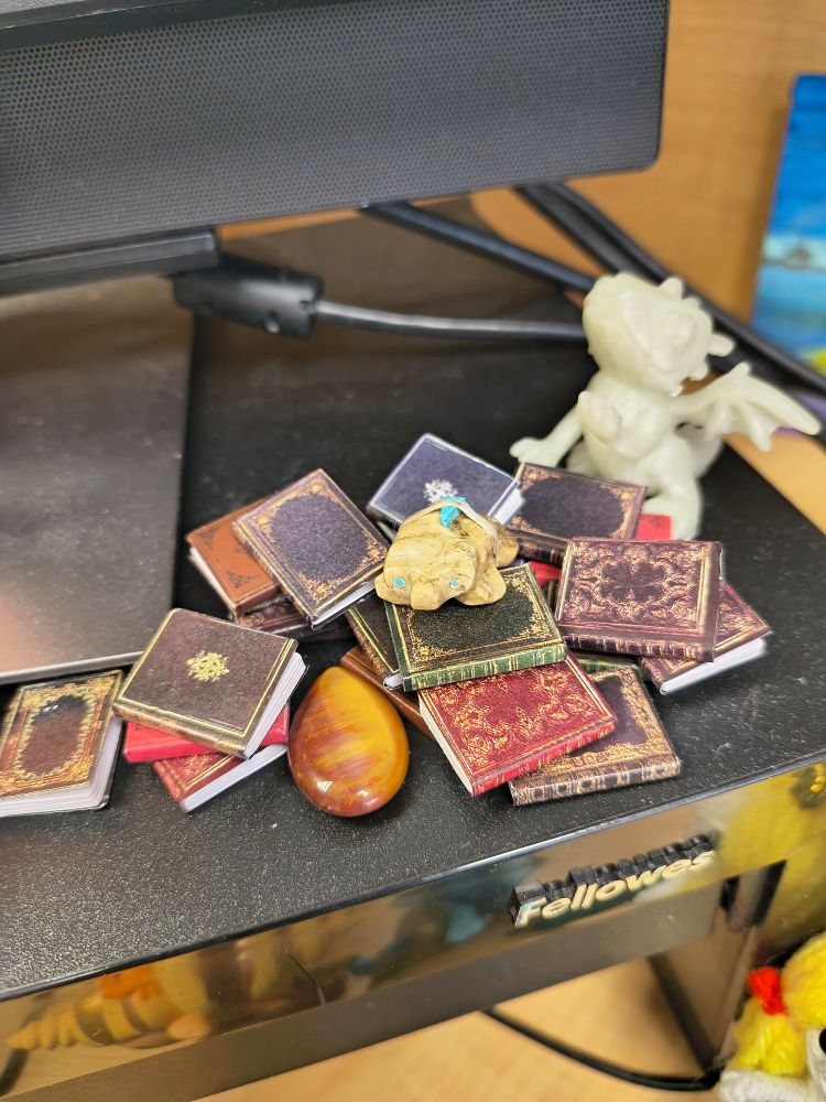 A small stone badger sits on collection of ornate miniature books stacked on a desk next to a small dragon figurine and a polished Tigers Eye stone. The scene feels whimsical and magical.