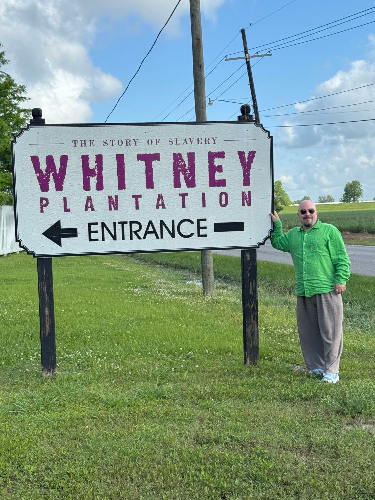 Me, bald with sunglasses, in a green shirt standing next to a sign that reads “Whitney Plantation” 