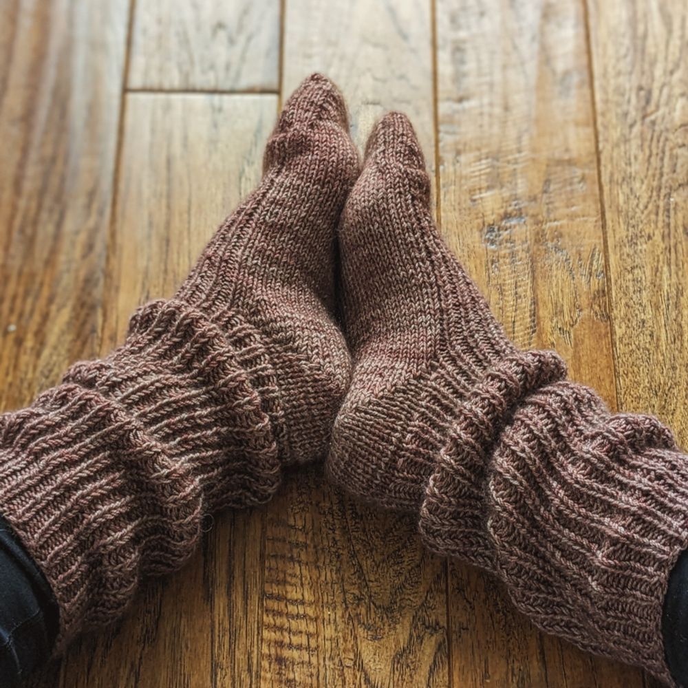 Women's feet wearing light plum colored socks. The leg of the socks have a slouchy ribbing on the leg. The pattern is Nest Socks by Katie Herrod of Songbird Handmade. The yarn is Yak and Merino Sock by Dark Lake Fiber Art.