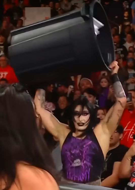 Rhea Ripley (center frame, focus) reeling back overhead a black plastic trash can filled with random, metallic junk to throw directly at Raquel Rodriguez (left corner, foreground) facing towards her, away from camera. #QueenShit #HaveAGoAfterIyo