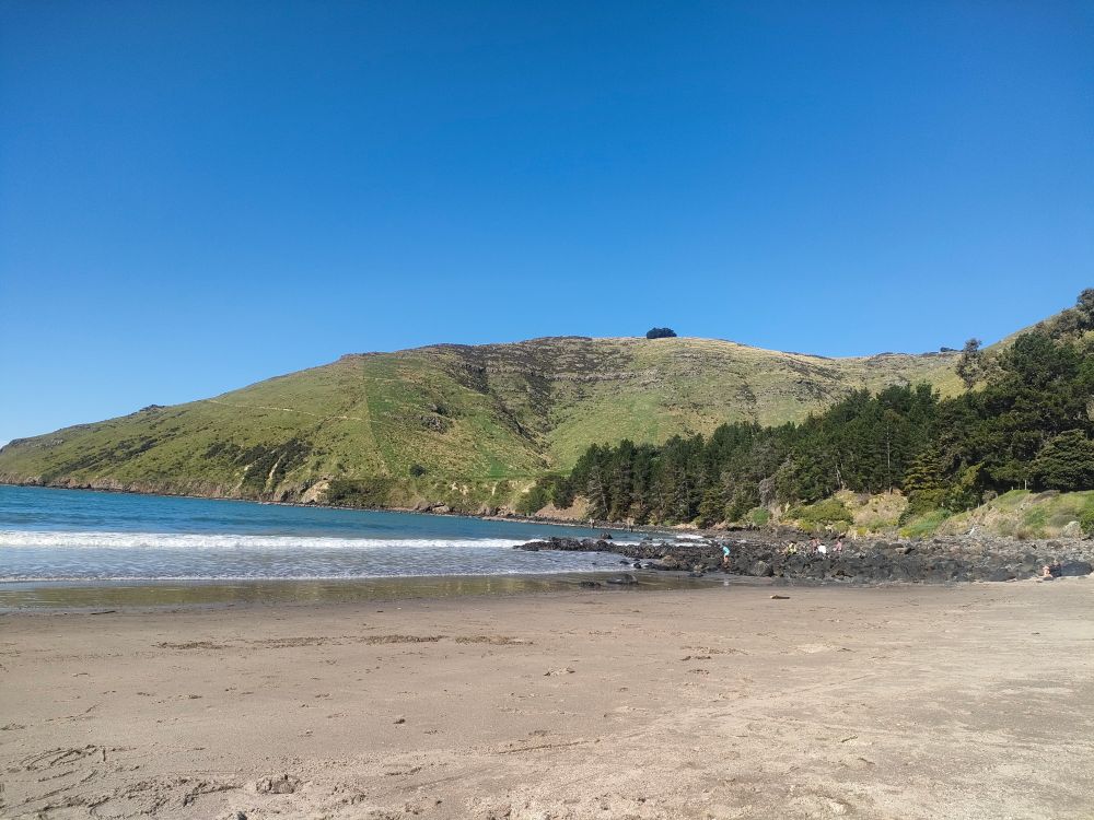 An empty beach in the foreground and about 6 people on some rocks. Flat blue sea to the left and green hills at the back