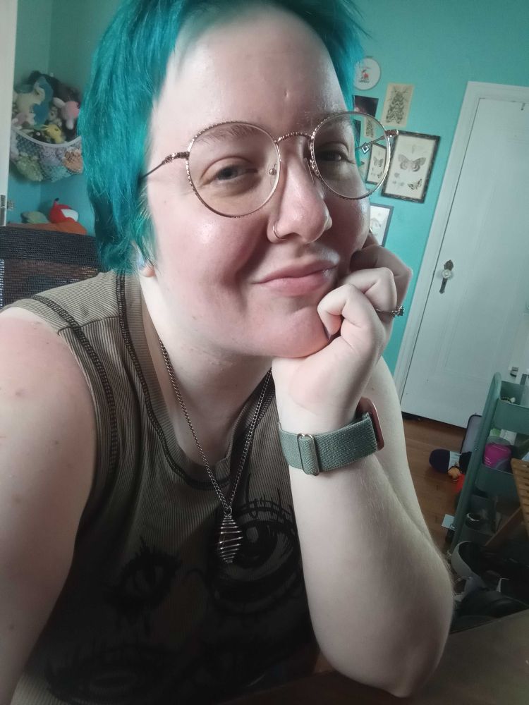 Image of Fox, a white person with blue hair and gold wire-frame glasses. They wear a green shirt and a crystal necklace and smile slightly at the camera.