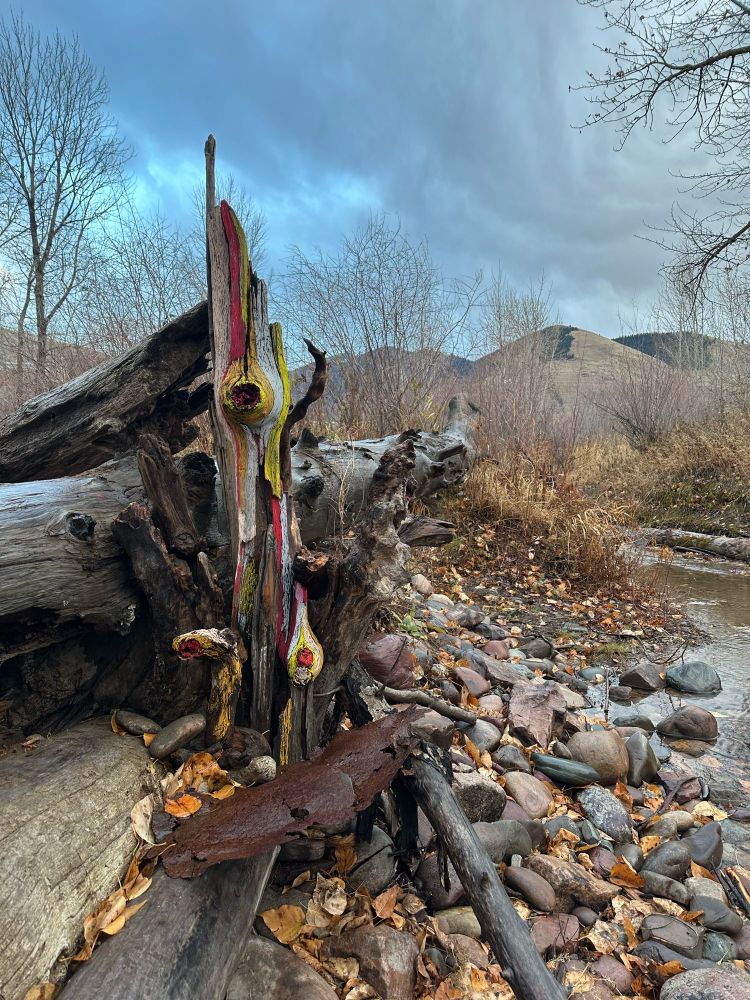 A bunch of logs and other detritus piled up near a stream but one of the pieces of driftwood has been painted in red, yellow-green, and light blue. The painting is very organic, following the natural lines of the wood. It has been propped so that the wood is vertical. 